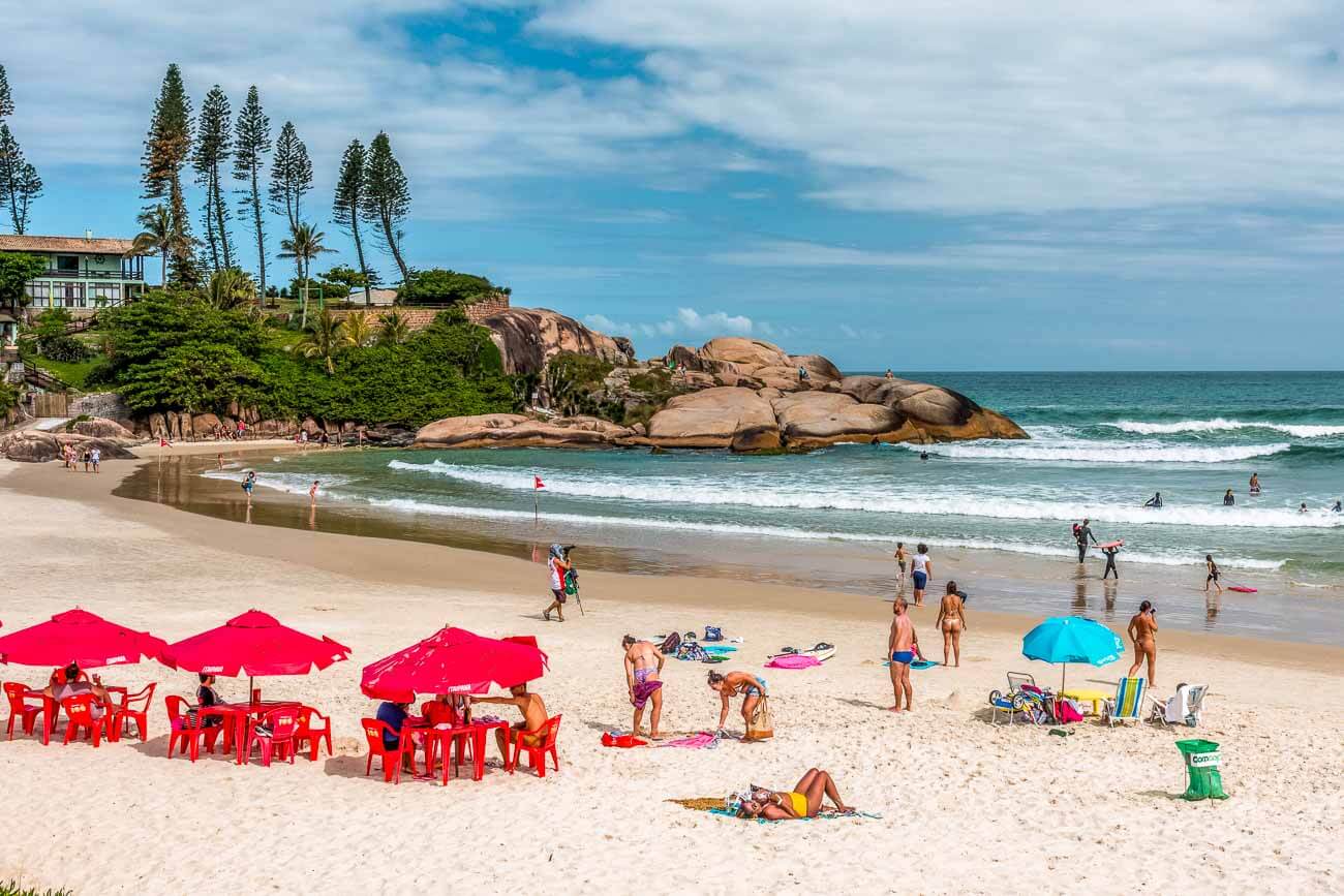 Mesas com cadeiras e guarda-sóis na Praia da Joaquina com algumas pessoas na areia e na água