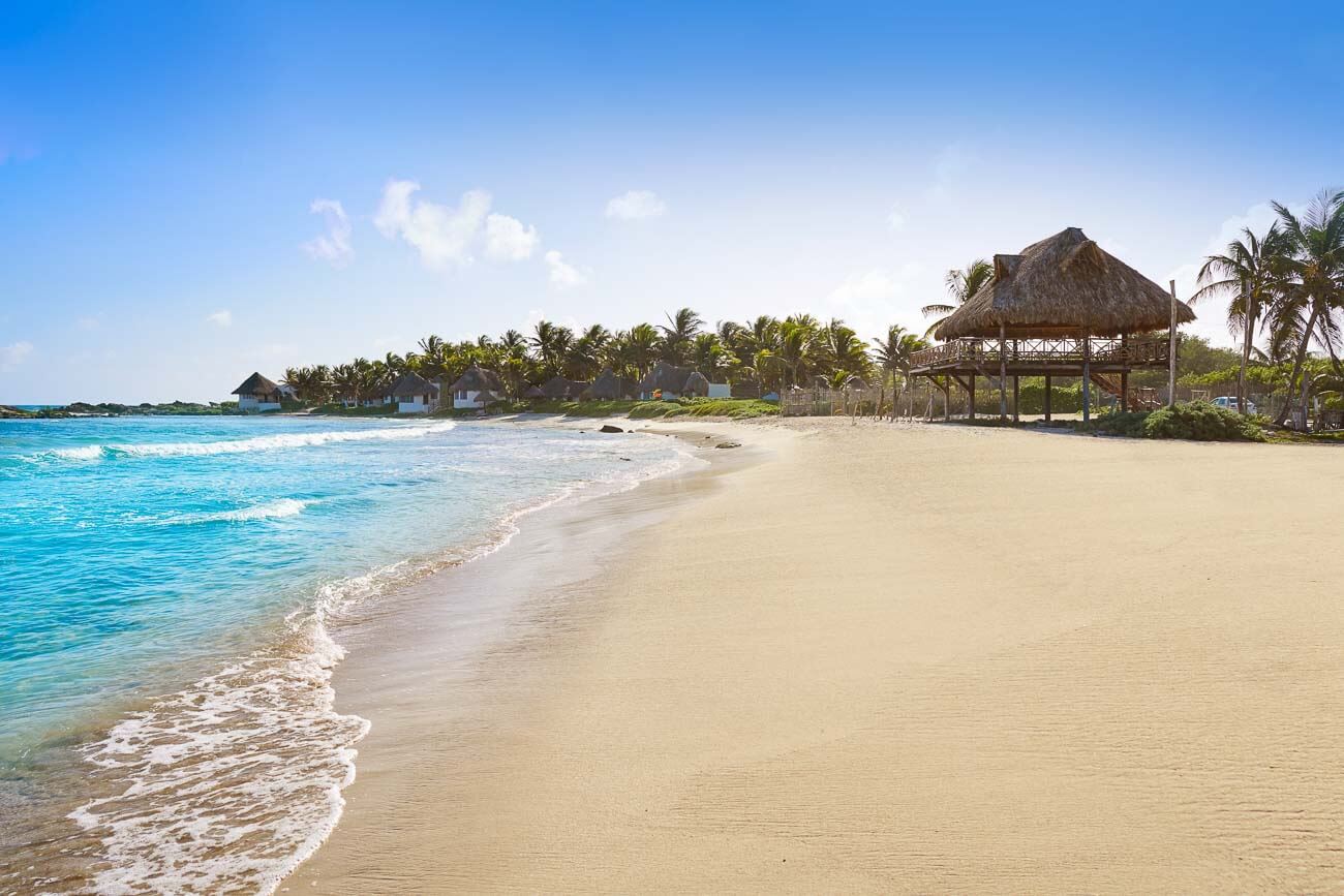 Faixa de areia larga e extensa em Praia de Tulum. A orla está repleta de coqueiros e construção rústica.