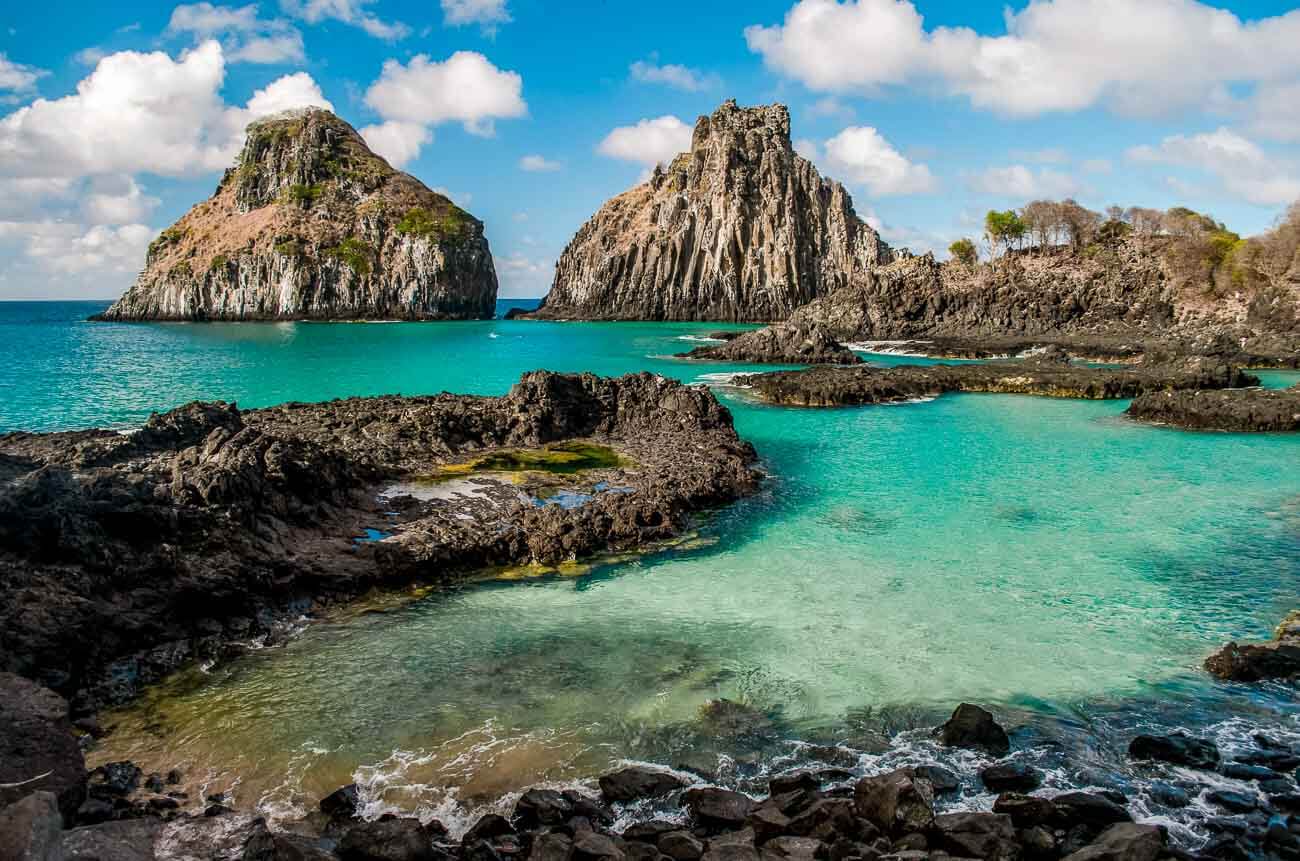Baía dos Porcos com o mar azul turquesa cristalino e formação de piscinas naturais. Ao fundo, o Morro Dois Irmãos