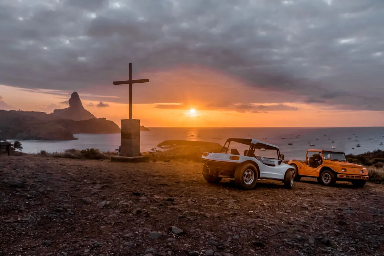 Dois buggys estacionados no alto de um morro com uma cruz em Fernando de Noronha durante o pôr do sol.