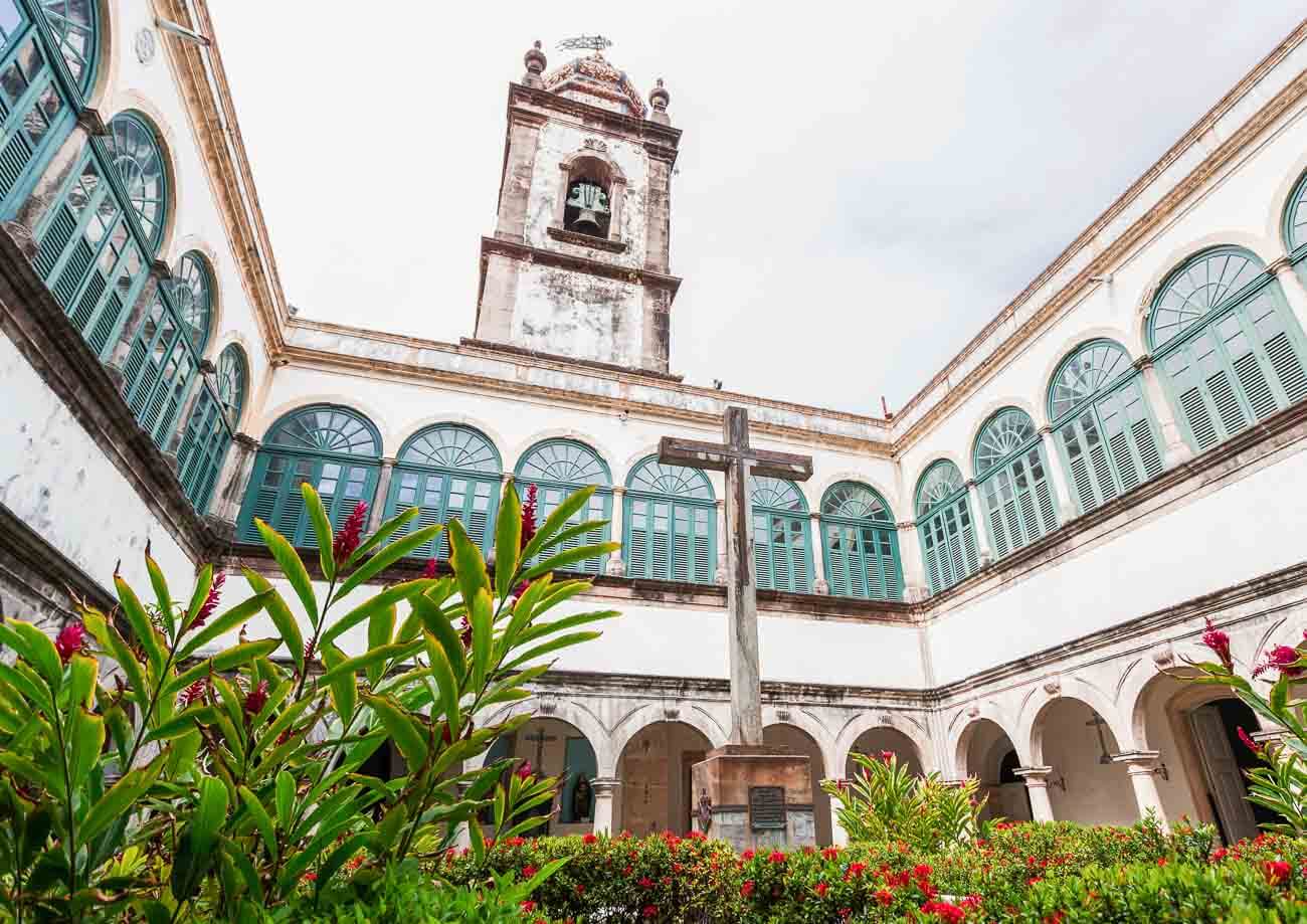 Área interna da Capela Dourada em Recife com um jardim ao ar livre e uma cruz;.