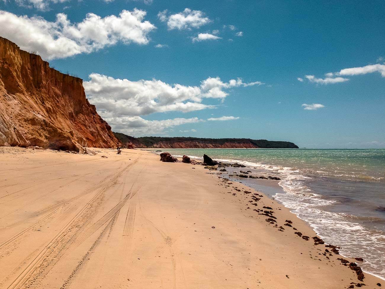 Faixa de areia vazia da Praia de Carro Quebrado em Alagoas com o mar sem ondas cristalinos e a orla repleta de falésias.
