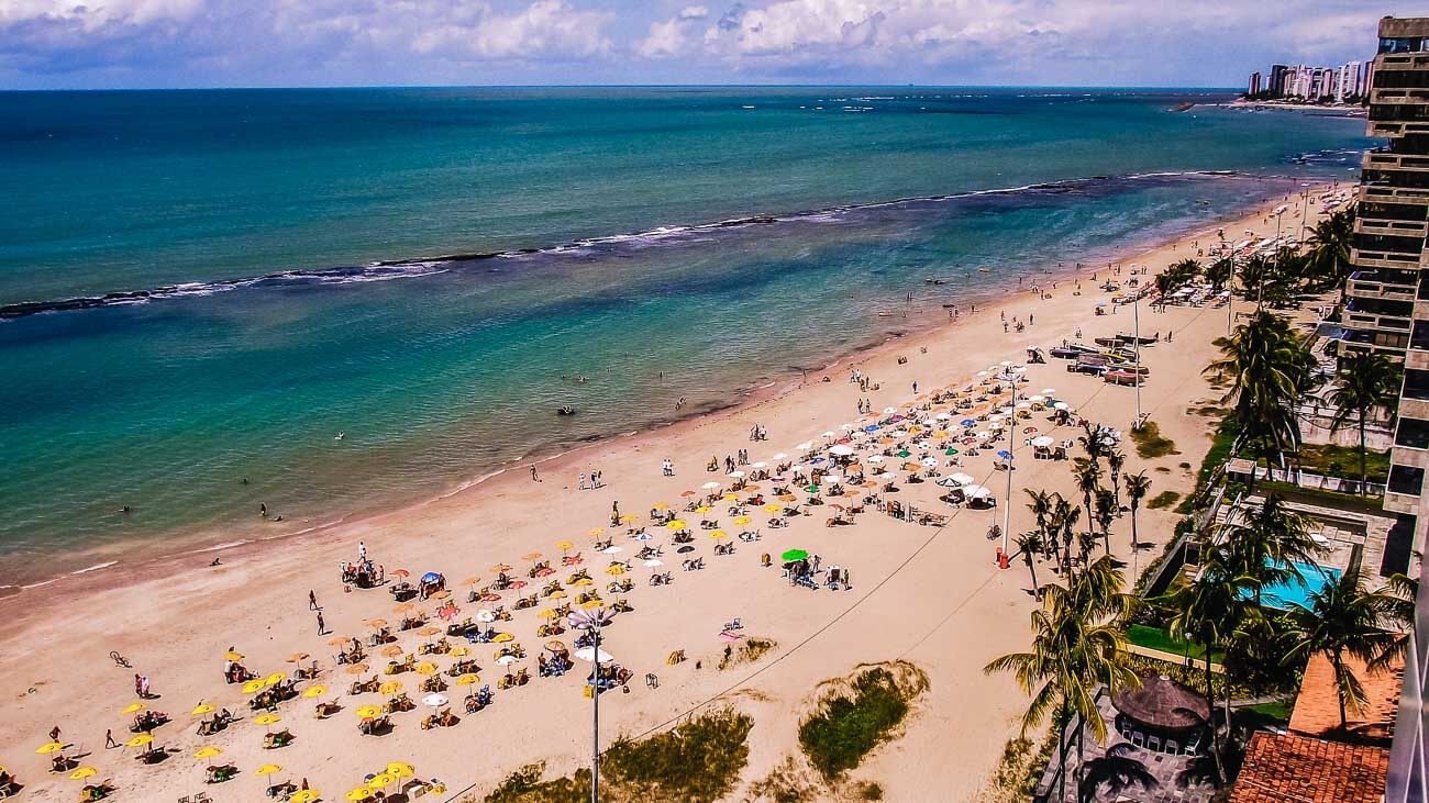 Vista de cima da Praia de Piedade, em Recife. A orla está repleta de guarda-sóis com mesas e cadeiras. O mar tranquilo sem ondas.