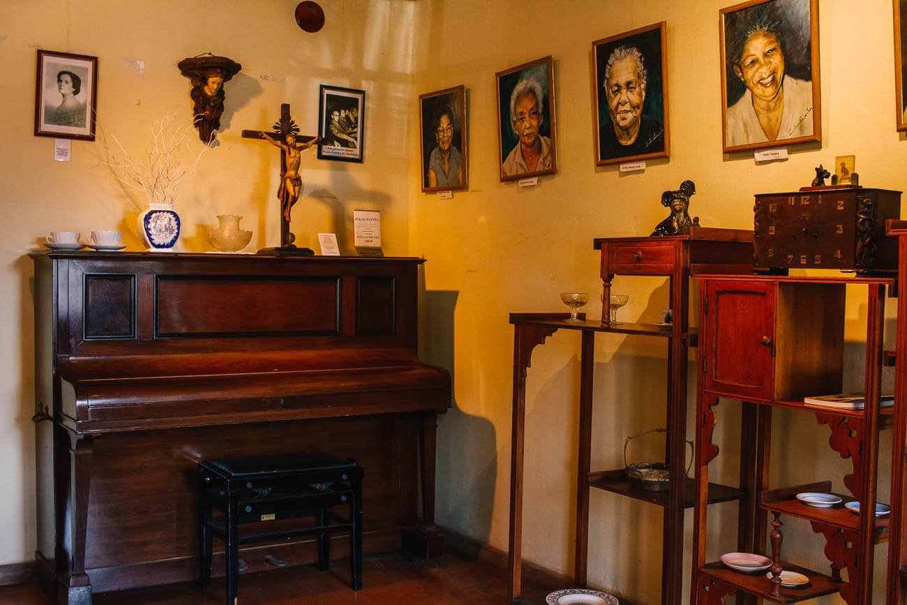 Área com um piano antigo e outros móveis de época e de madeira, com quadros de rostos de pessoas na parede na Casa de Cultura Doutor Bento Costa Junior.