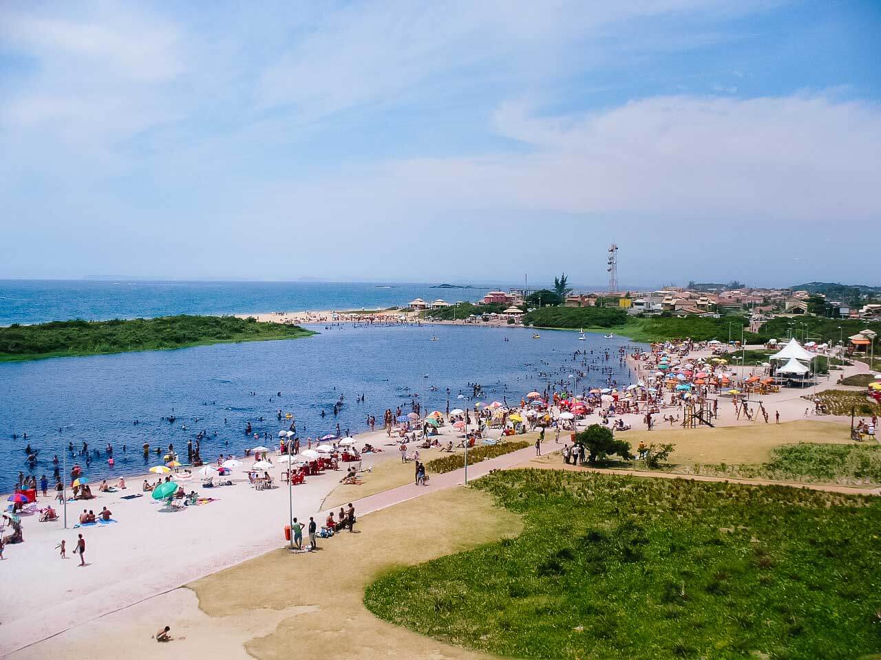Vista da Lagoa do Iriri em Rio das Ostras, com várias pessoas na água e nas margens, com guarda-sóis, cangas e cadeiras.