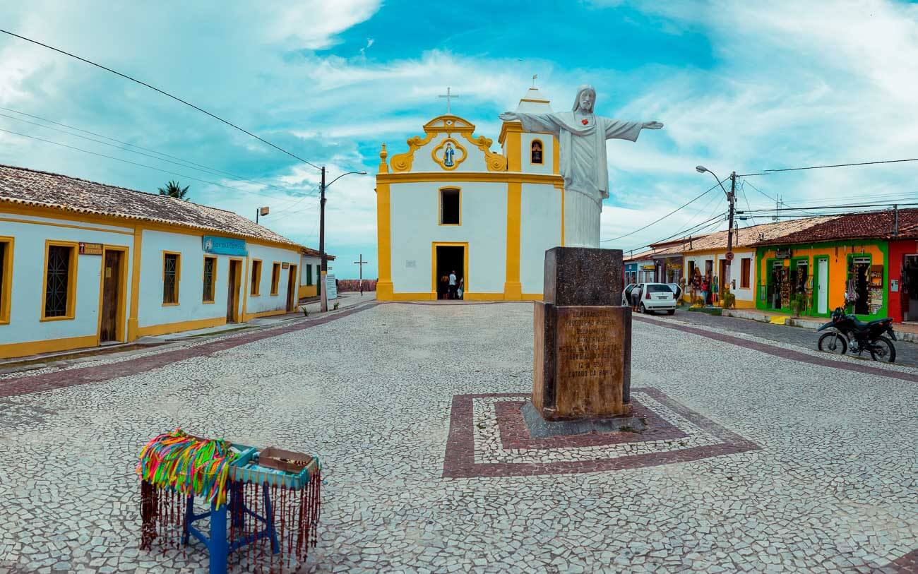 Fachada da Igreja Nossa Senhora d'Ajuda com uma escultura de Jesus Cristo na praça em frente.