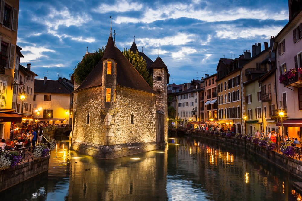Rio Thiou, em Annecy, iluminado por luzes amarelas e artificiais ao entardecer, em Annecy. Há pessoas em frente a restaurantes às margens do rio.