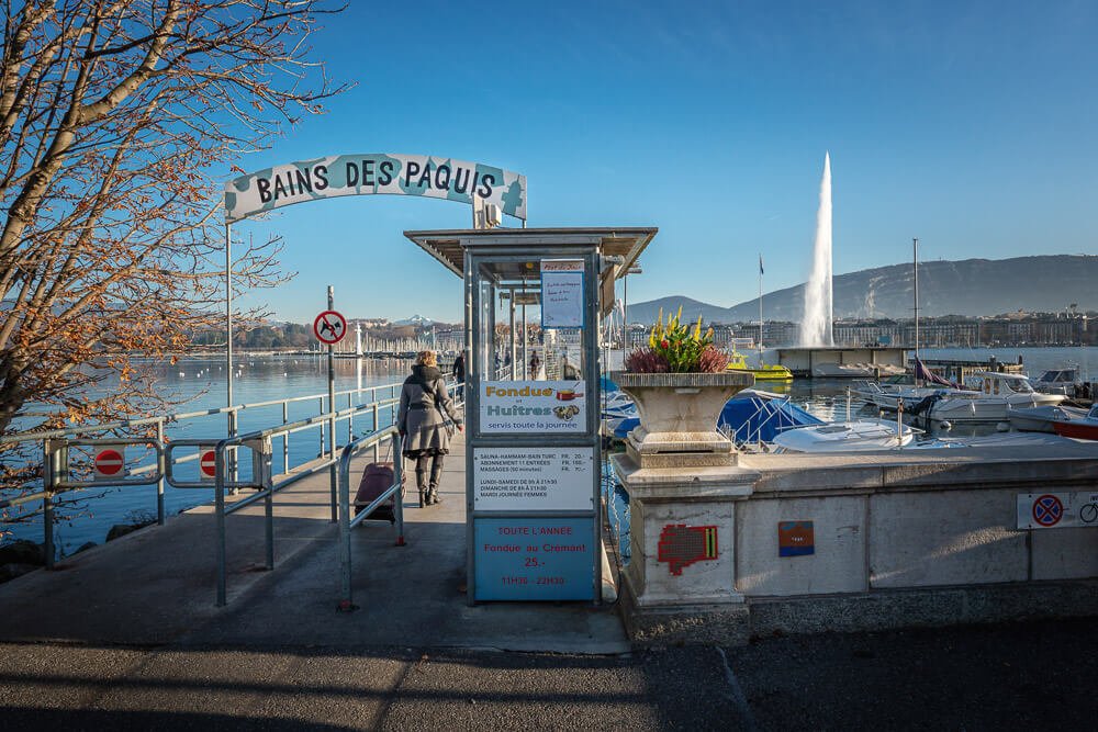 Entrada do Bains de Pâquis, à beira do Lago Léman, em Genebra. Há uma passarela em concreto, barcos ao fundo e anúncios de fondue.