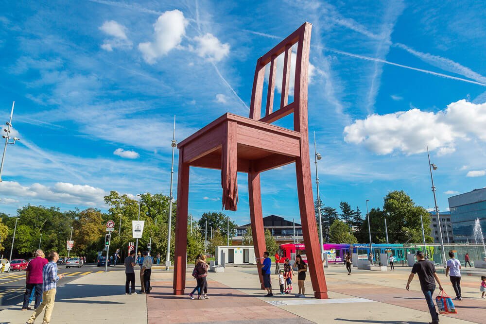 Grande escultura Cadeira Quebrada, em Genebra. Há uma escultura enorme de uma cadeira com uma das pernas quebrada. Há pessoas em volta da atração.