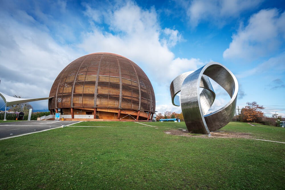 Visão externa do moderno prédio do CERN, em Genebra. À esquerda, há um prédio em formato de semiesfera e, em sua frente, uma contemporânea escultura feita com metal prateado.
