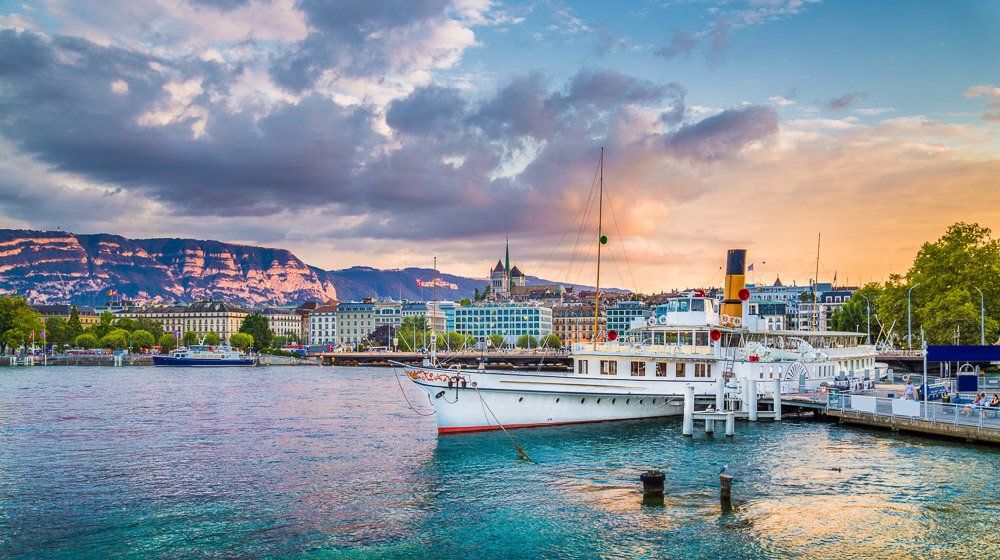 Parte do Lago Léman nas proximidades da região da Cidade Velha, em Genebra. Há um barco branco em destaque, na lateral direita, e montanhas atrás de prédios, na lateral esquerda.
