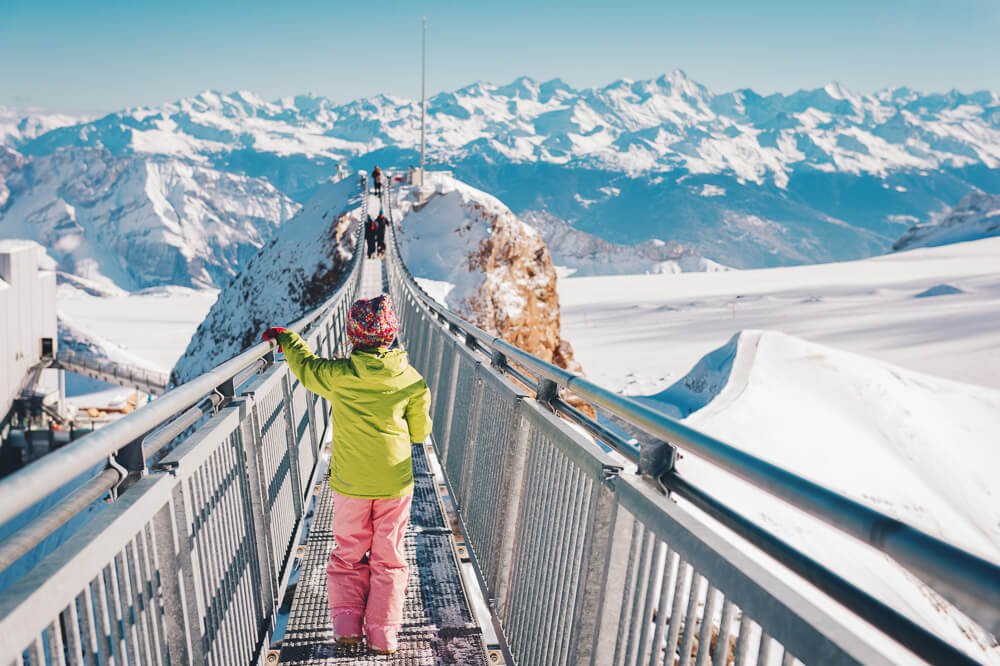 Ponte Suspensa sobre os alpes suíços cobertos pela neve. Há pessoas atravessando a ponte.