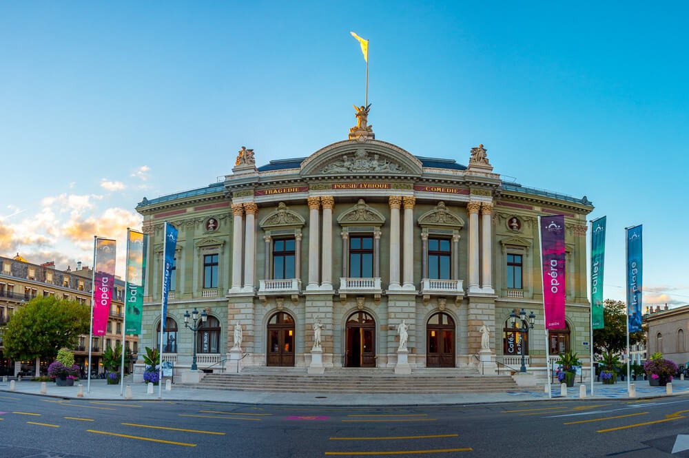 Parte frontal do Grand Théâtre de Genève. Há uma edifício com arquitetura clássica, oito colunas, três portas de madeira e paredes em tons claros.
