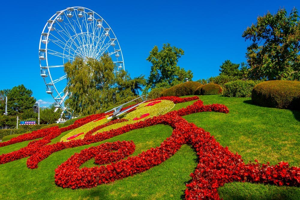 Relógio paisagístico feito de flores no Jardin Anglais. Há flores vermelhas e folhas verdes em um gramado inclinado, formando um relógio. Há também uma roda gigante à esquerda.