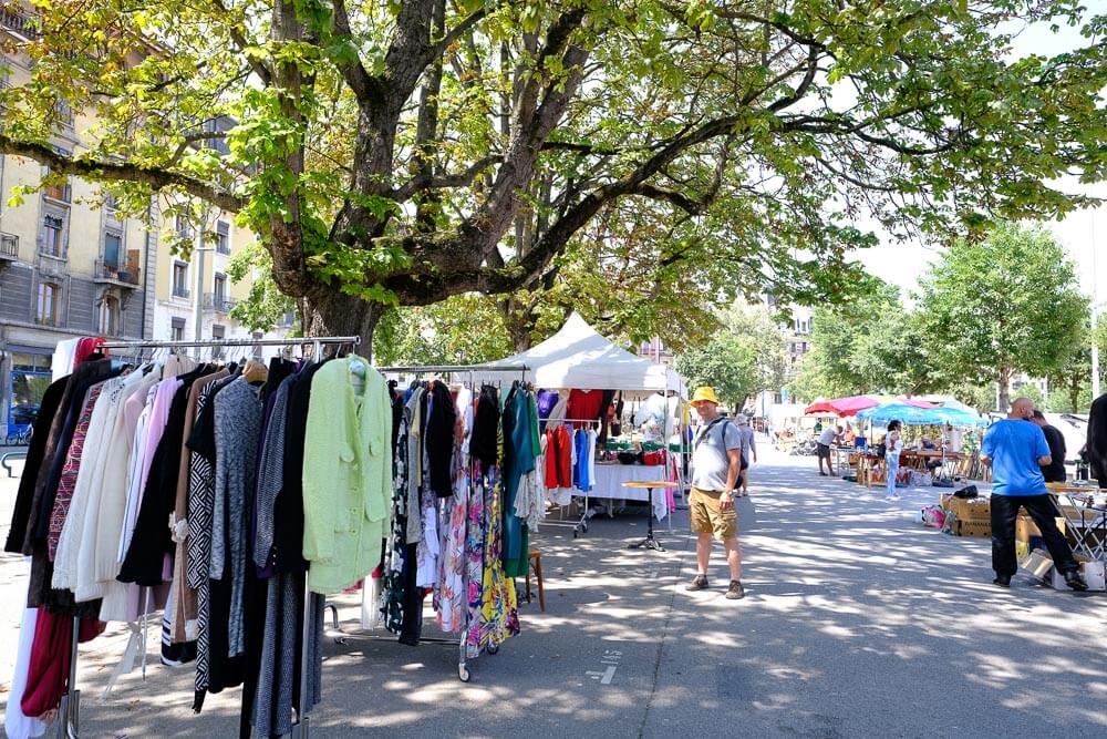 Mercado de Pulgas, em Genebra. Há um comércio ao ar livre com roupas em araras, pessoas comprando e vendendo sob as árvores do local.