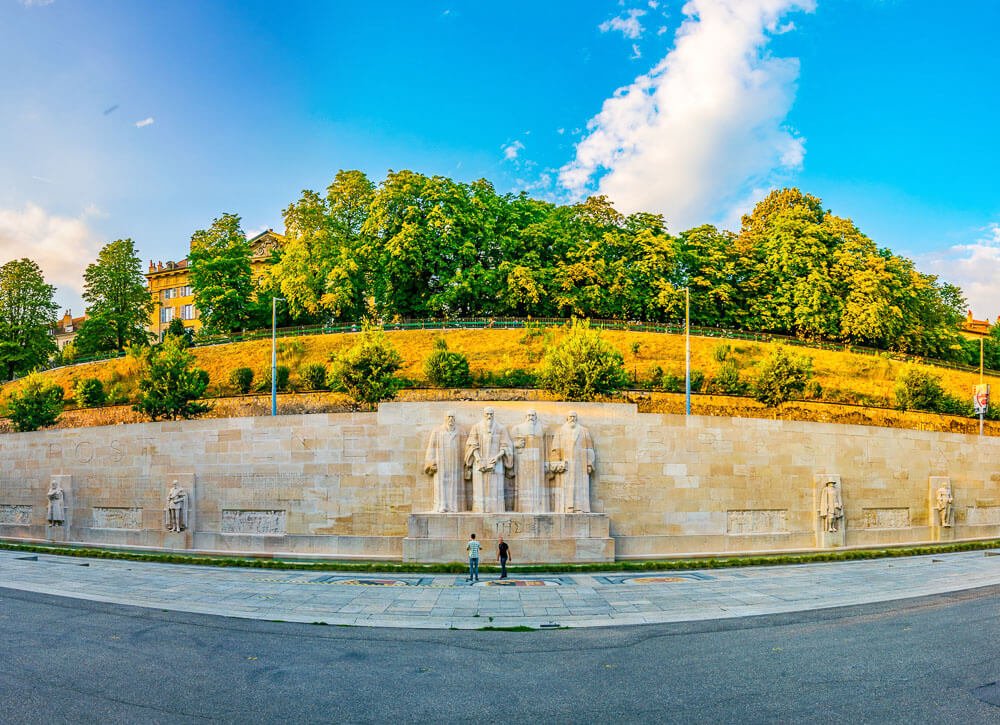 Fachada do Muro dos Reformadores, feito de pedras claras, com quatro grandes estátuas principais ao meio e mais quatro distribuídas nas laterais. Há um jardim acima do muro, árvores no topo e duas pessoas próximas às estátuas.