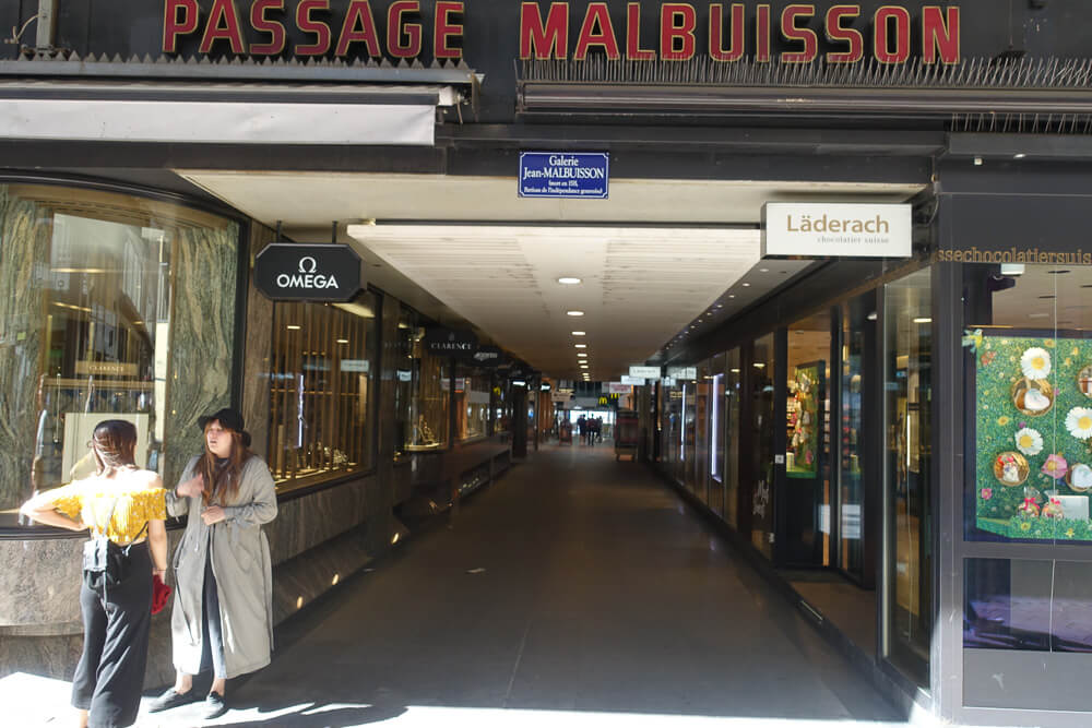 Hall de entrada da Passage Malbuisson, em Genebra. Há um corredor com estabelecimentos nas laterais e duas pessoas na entrada.