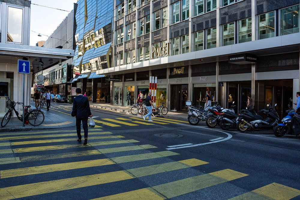 Rue du Rhône em um fim de tarde. Há pessoas circulando e motos estacionadas à direita.