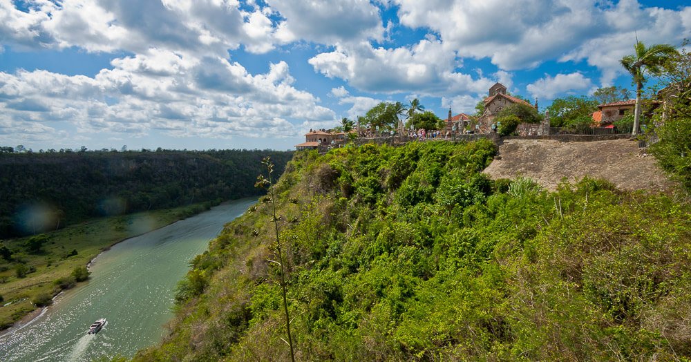 Construções do vilarejo dos Altos de Chavón, em La Romana, sobre morro coberto de vegetação, cercado pelo rio Chavón, sob um céu azul com diversas nuvens.