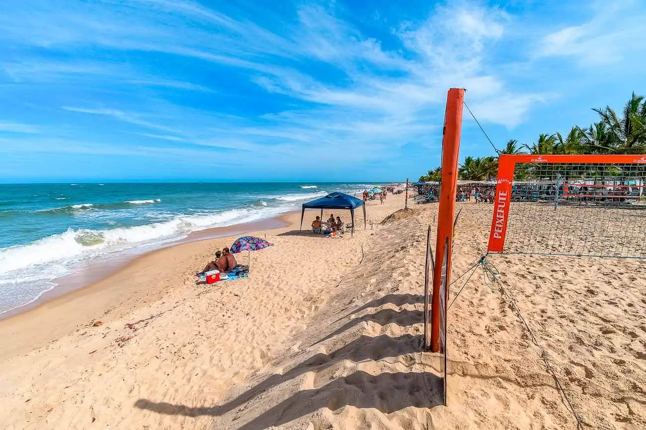 Faixa de areia da Praia dos Nativos com rede de vôlei e pessoas na areia.