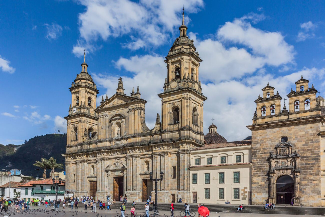 Pessoas transitando em várias direções, em frente à escadaria cinza da catedral basílica metropolitana de Bogotá, em tons terrosos.