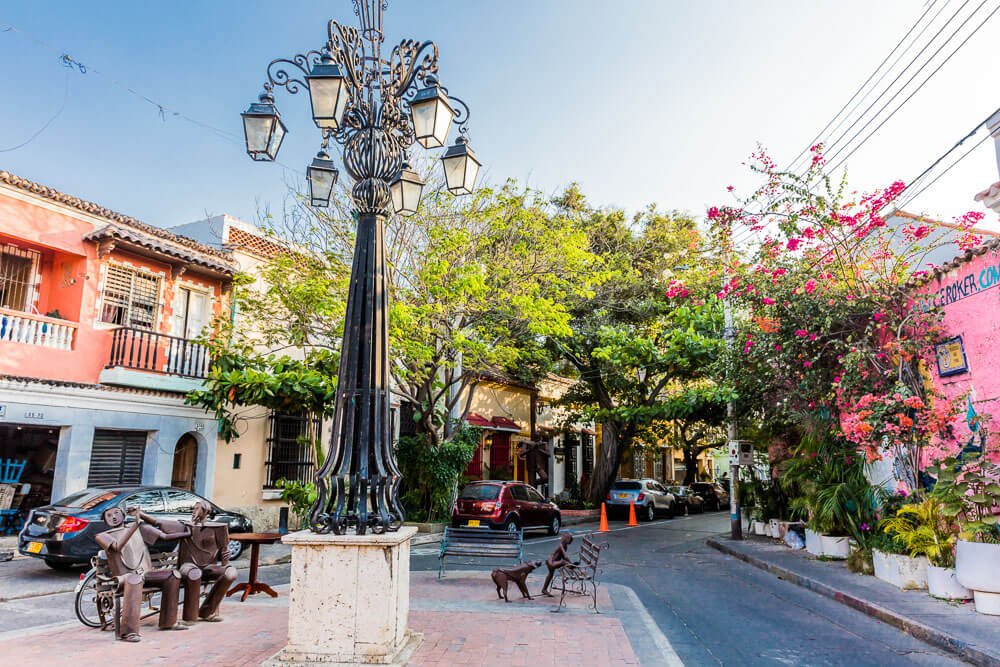 Rua no bairro de Getsemaní, em Cartagena. Há muitas árvores, flores e alguns carros circulando pela rua embelezada pela arquitetura colonial presente na cidade.