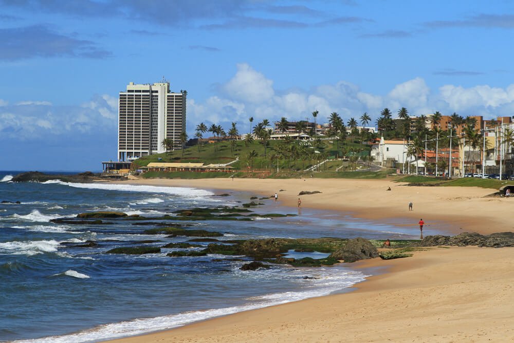 Praia de Amaralina com mar de águas agitadas, um prédio à beira-mar e vegetações litorâneas.