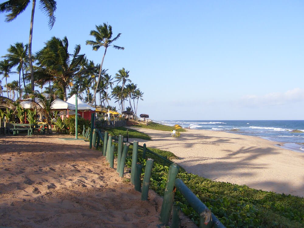 Praia de Stella Maris com extensa faia de areia, coqueiros e barracas em frente ao mar.