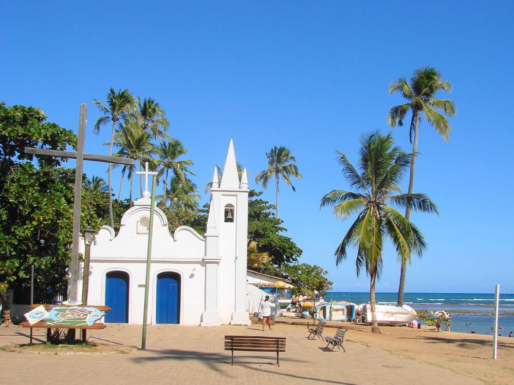 Praia do Forte com areia clara, pequena igreja com fachada branca, coqueiros e mar na lateral direita.