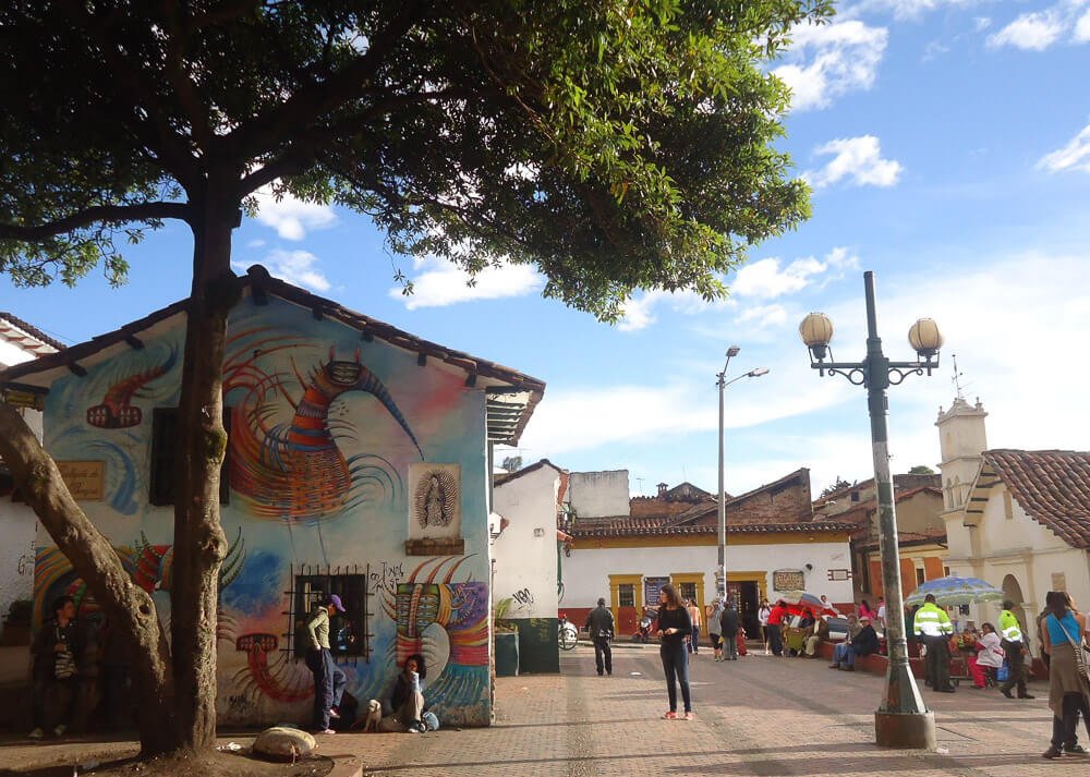 Rua colorida na região de La Candelaria. Há pessoas, igreja e uma árvore no local.