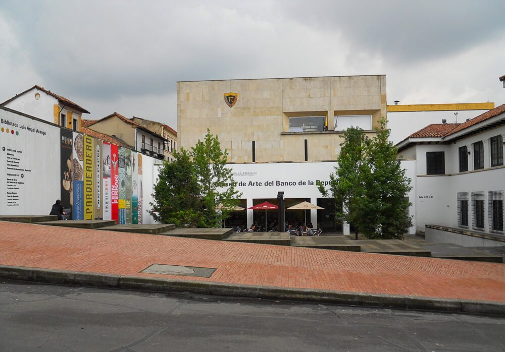Colección de Arte de Banco de la República com árvores, calçada avermelhada e pessoas sentadas em cadeiras com mesas.