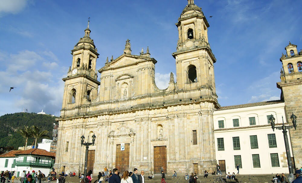 Praça com catedral no centro. Há pessoas em frente a catedral com fachada bege.