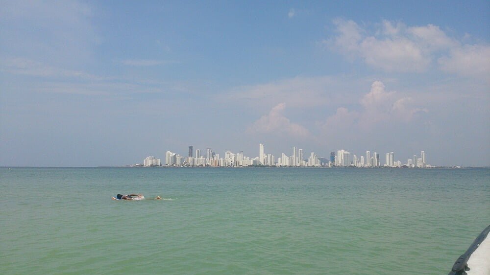 Litoral da ilha de Tierra Bomba. Há prédios brancos ao fundo e uma pessoa nadando no meio do mar de águas azuis-turquesa.