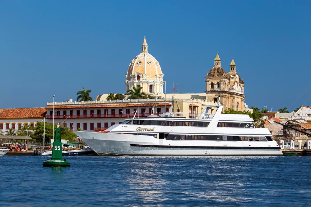Embarcação de médio porte sobre o mar azul em um dia de sol. Ao fundo, as construções tradicionais da cidade de Cartagena.