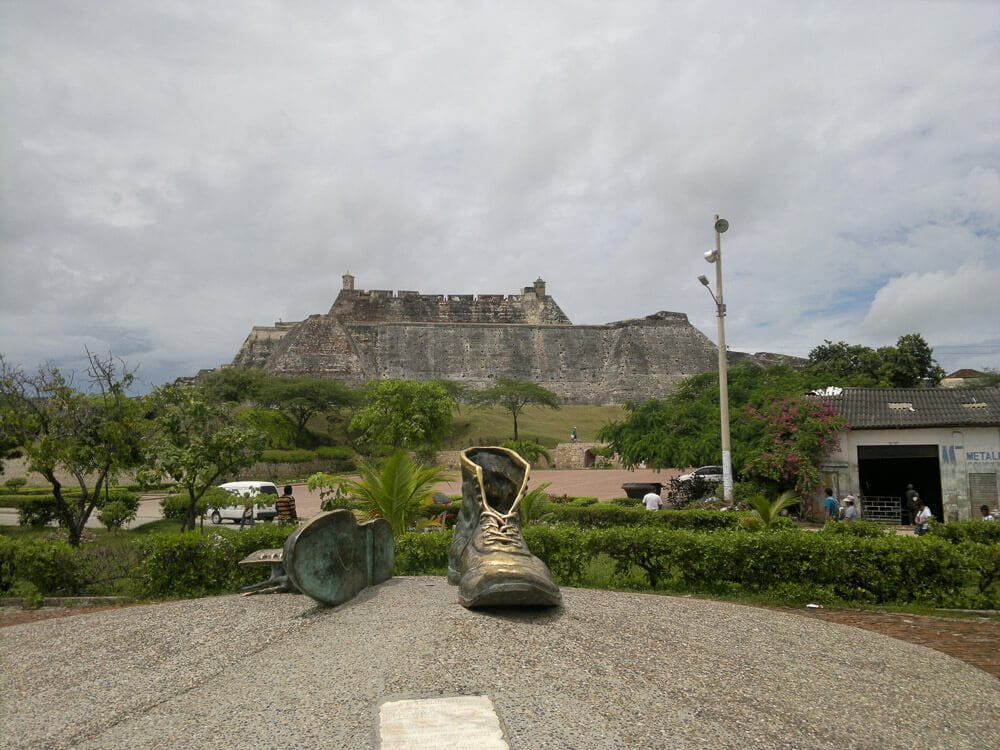 Escultura de dois sapatos cinzas com fortaleza ao fundo da paisagem.