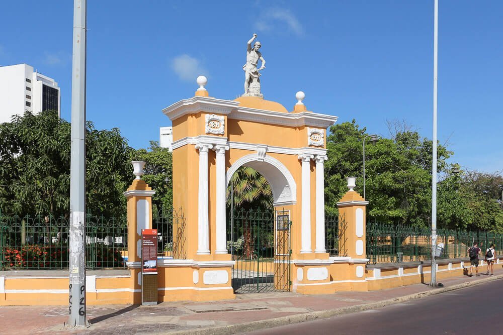 Fachada laranja do Parque del Centenario. Há muitas árvores atrás do pórtico e um portão de grades.