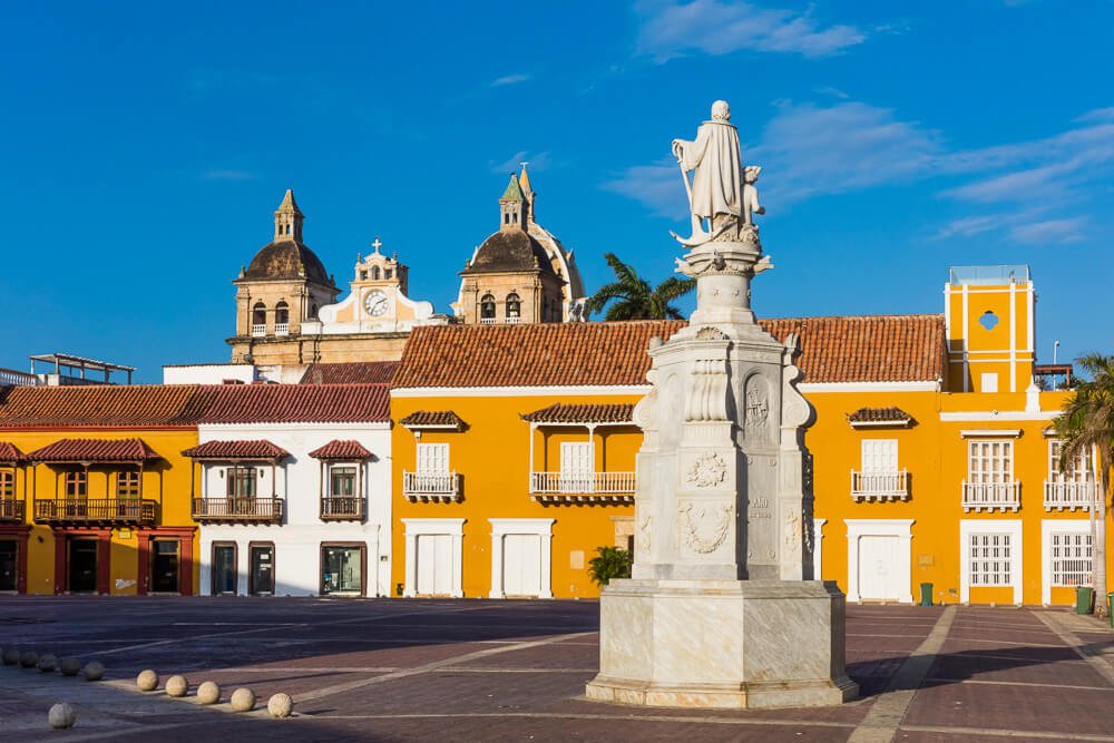 Estátua branca no meio da Plaza de la Aduana. Ao fundo, casas coloniais nas cores amarelo e branco.