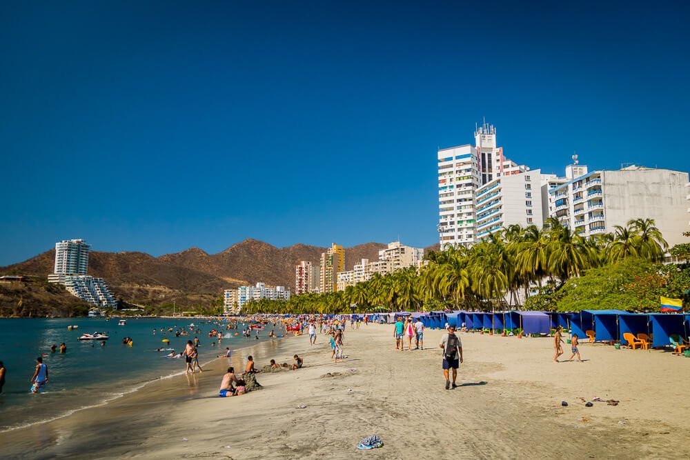 Praia em Santa Marta. À esquerda há uma montanha marrom. Na lateral direita, há prédios à beira-mar.
