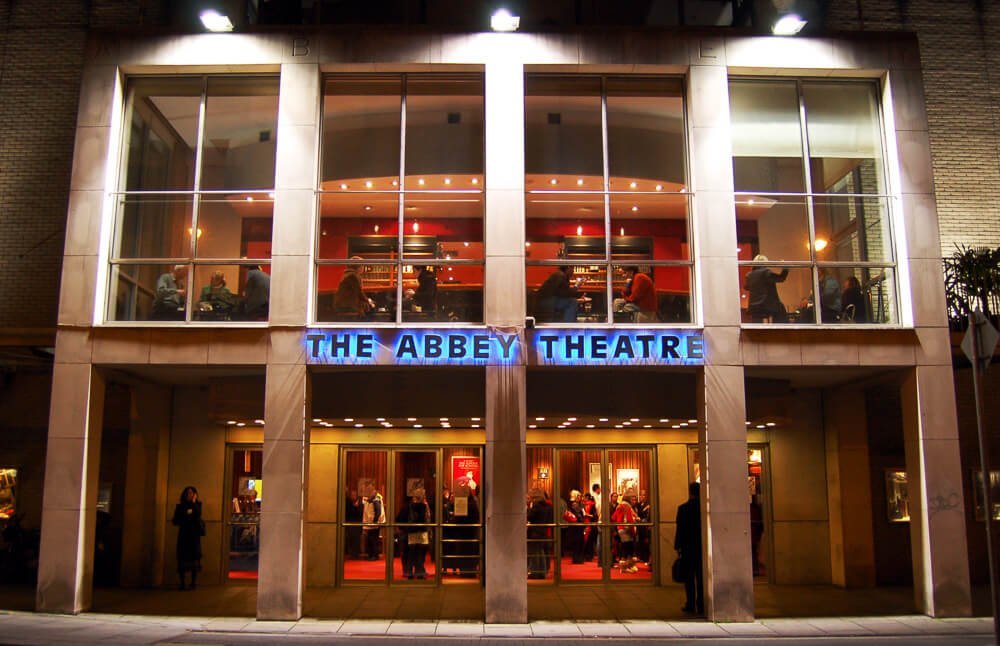 Fachada iluminada do Abbey Theatre. O prédio do teatro possui dois andares e janelas de vidro na fachada.