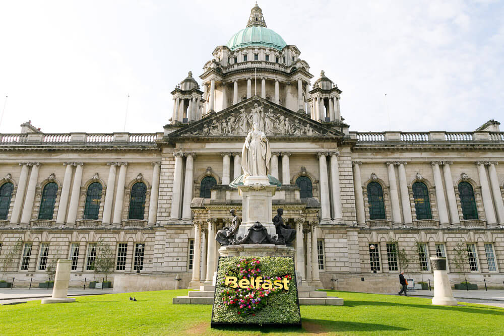 Entrada da prefeitura de Belfast. Há um prédio majestosos com muitas colunas, janelas e uma estátua no pátio em frente.