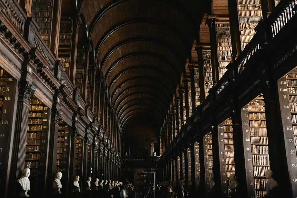 Interior da Biblioteca Long Room, na Trinity College Dublin. Há muitos livros com lombadas marrons dispostos em estantes altas e bustos nas laterais.