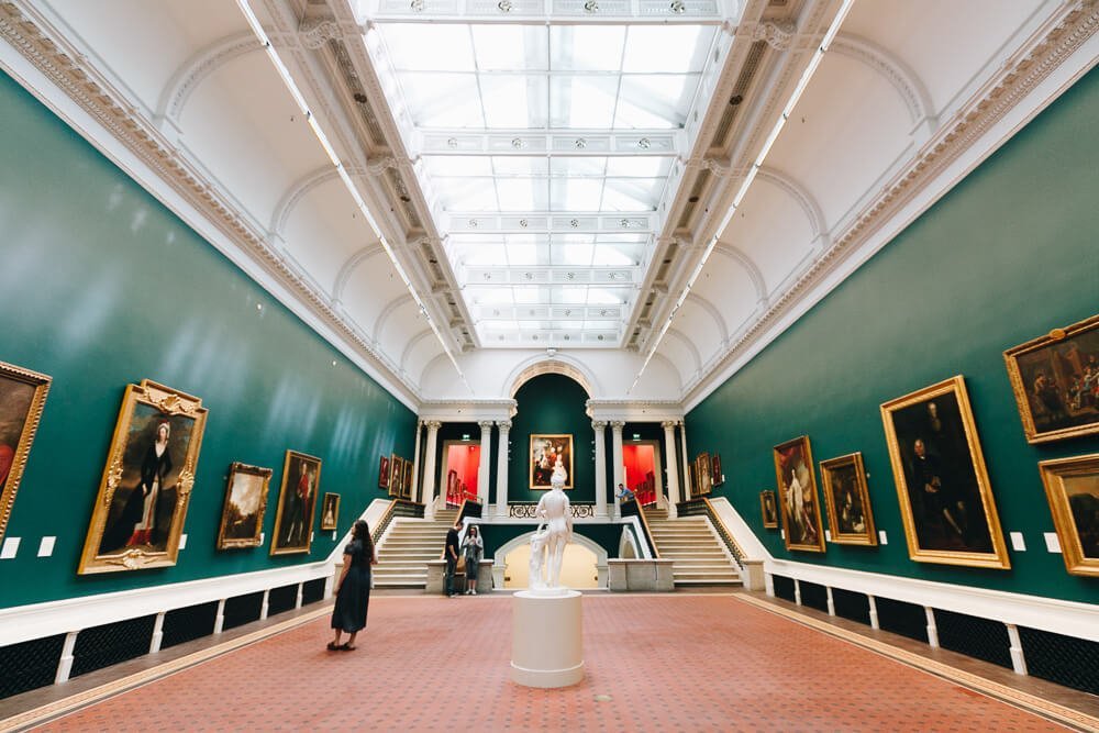 Interior da Galeria Nacional da Irlanda. Há pinturas nas paredes laterais e uma escultura no centro do ambiente.