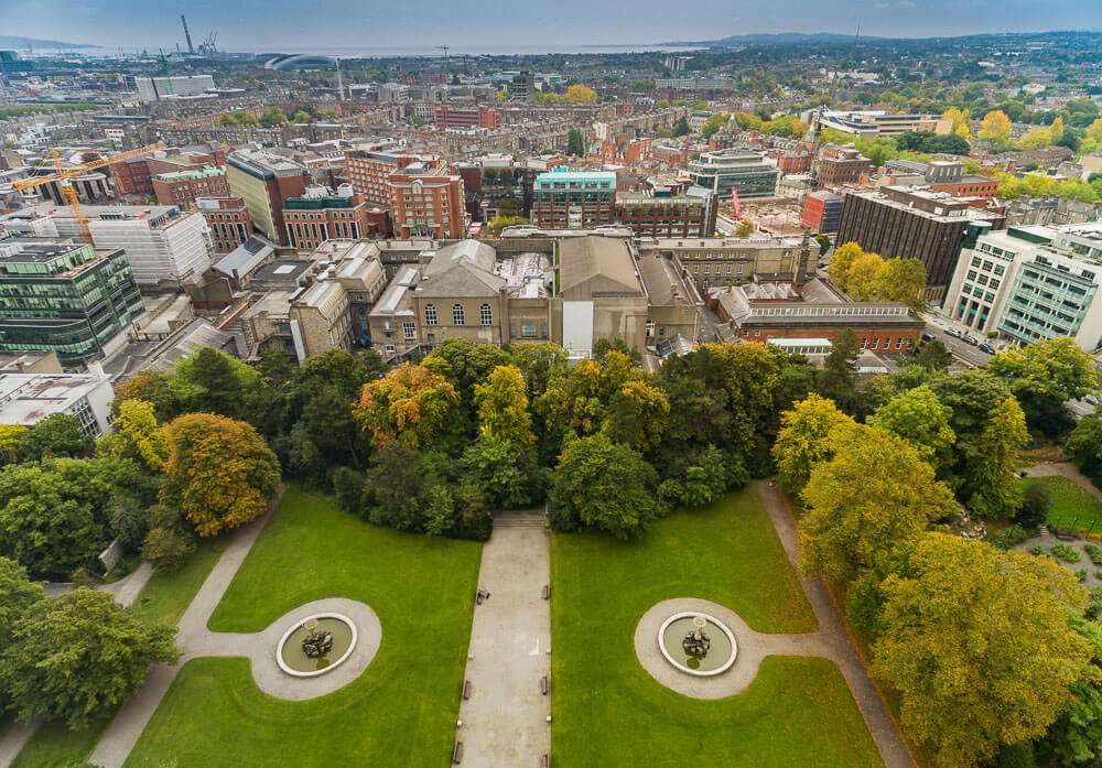 Visão aérea do Iveagh Gardens. Há um jardim no meio e, ao fundo, há os prédios do centro de Dublin.