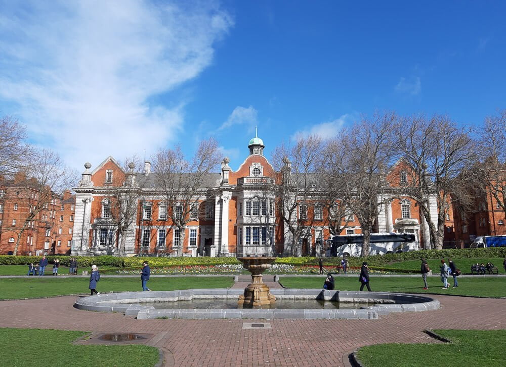 St. Patrick’s Park com árvores, uma fonte e jardins. Há fachadas de prédios aos fundos.