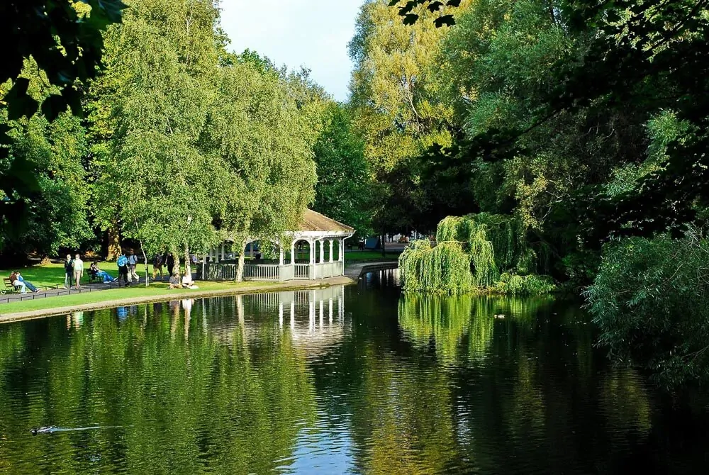 Parque Saint Stephen’s Green com lago refletindo as árvores ao redor. Há um coreto e pessoas caminhando à esquerda.