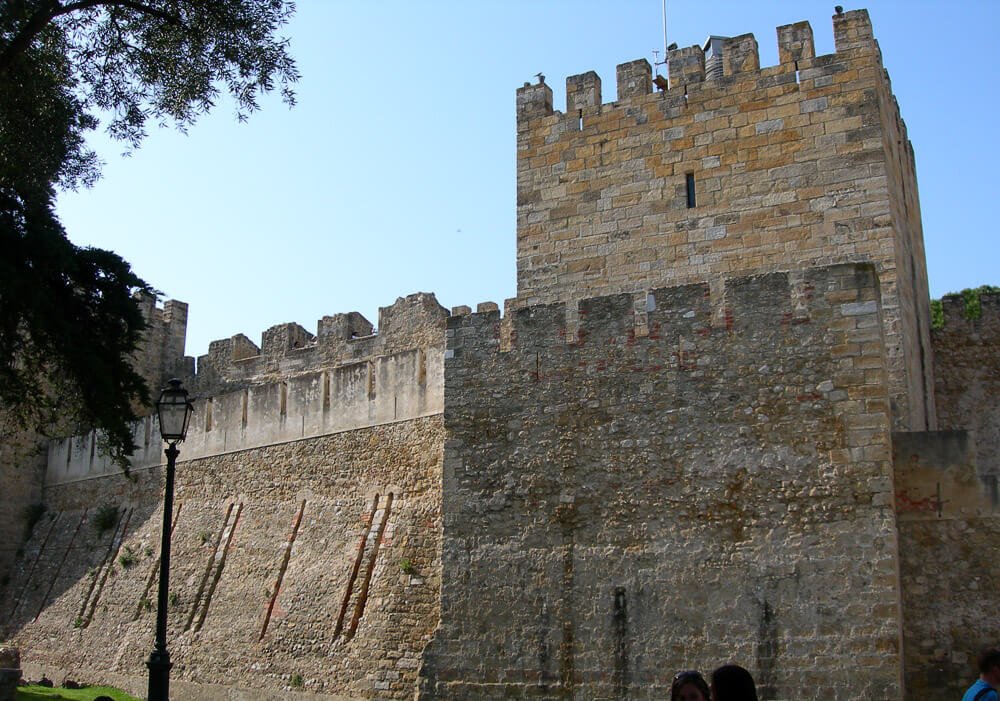 Castelo de São Jorge com paredes feitas de pedras claras, uma lâmpada antiga à esquerda e pessoa visitando o local.