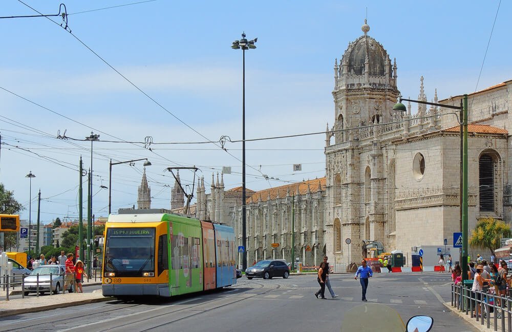 Rua em Lisboa com trem passando à esquerda e Mosteiro dos Jerónimos à direita.