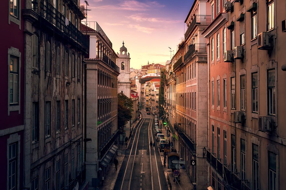 Entardecer na Rua de São Paulo, em Lisboa. Há prédios nas laterais da rua estreita.