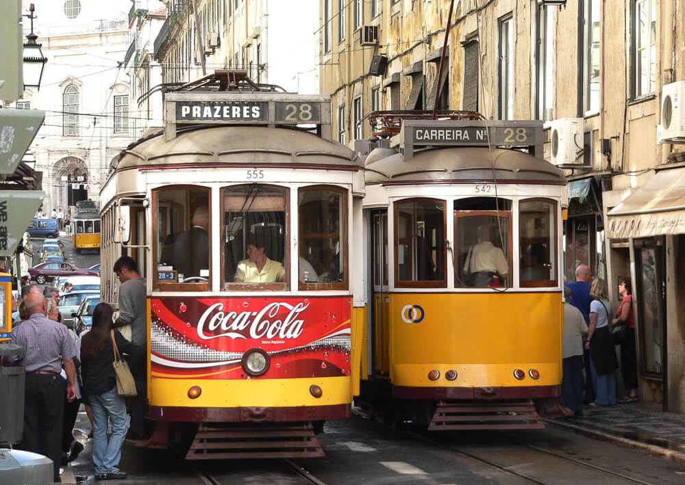 Dois bondinhos elétricos antigos de Lisboa nas cores amarelo, branco e marrom. O bondinho da esquerda tem uma propaganda da Coca-Cola na parte frontal. Na rua, há muitas pessoas, carros e prédios ao redor.