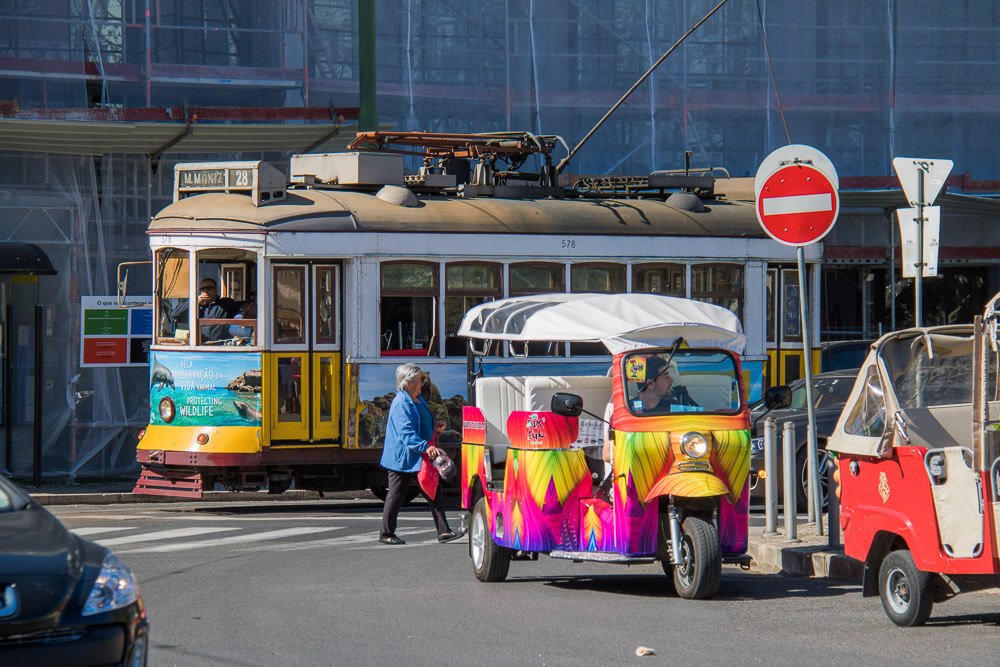 Rua com bonde elétrico e tuk-tuks estacionados à direita.